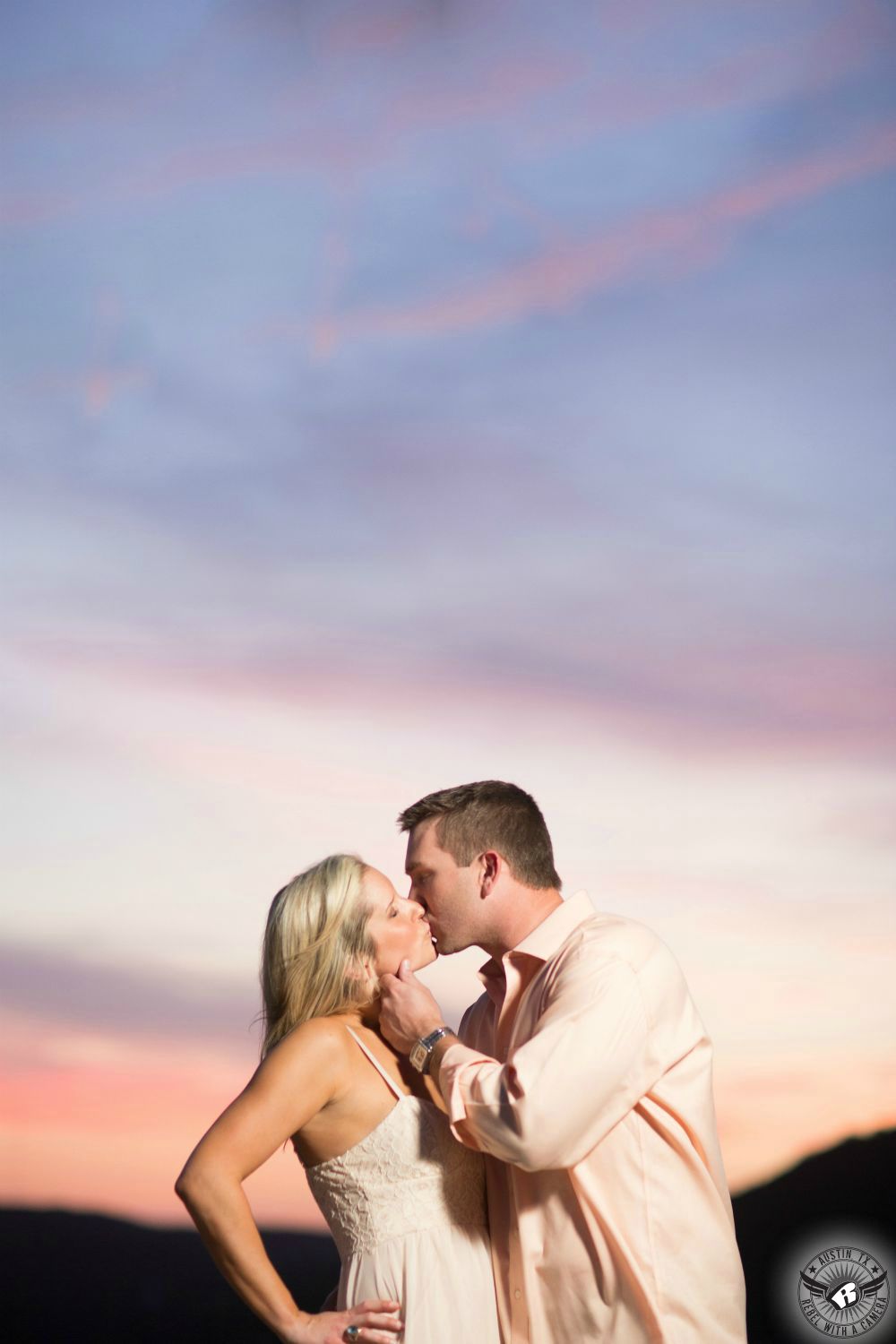 blond girl in a string tank top tenderly kisses a brunette guy in a pink button up shirt in front of a twilight sky willed with blue, purple and pink colors in this engagement photo from the pennybacker bridge in austin 